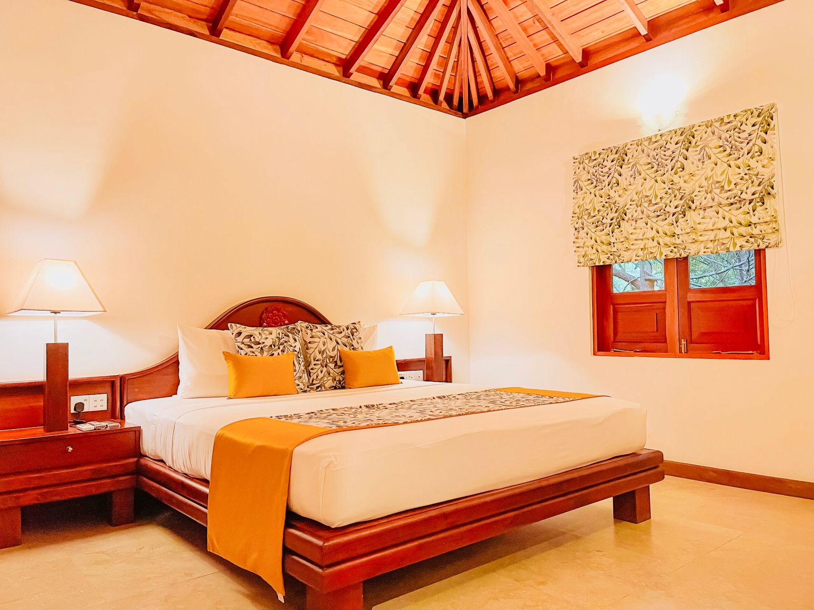 A villa bedroom under a pitched timber ceiling, with a carved headboard and an orange throw.